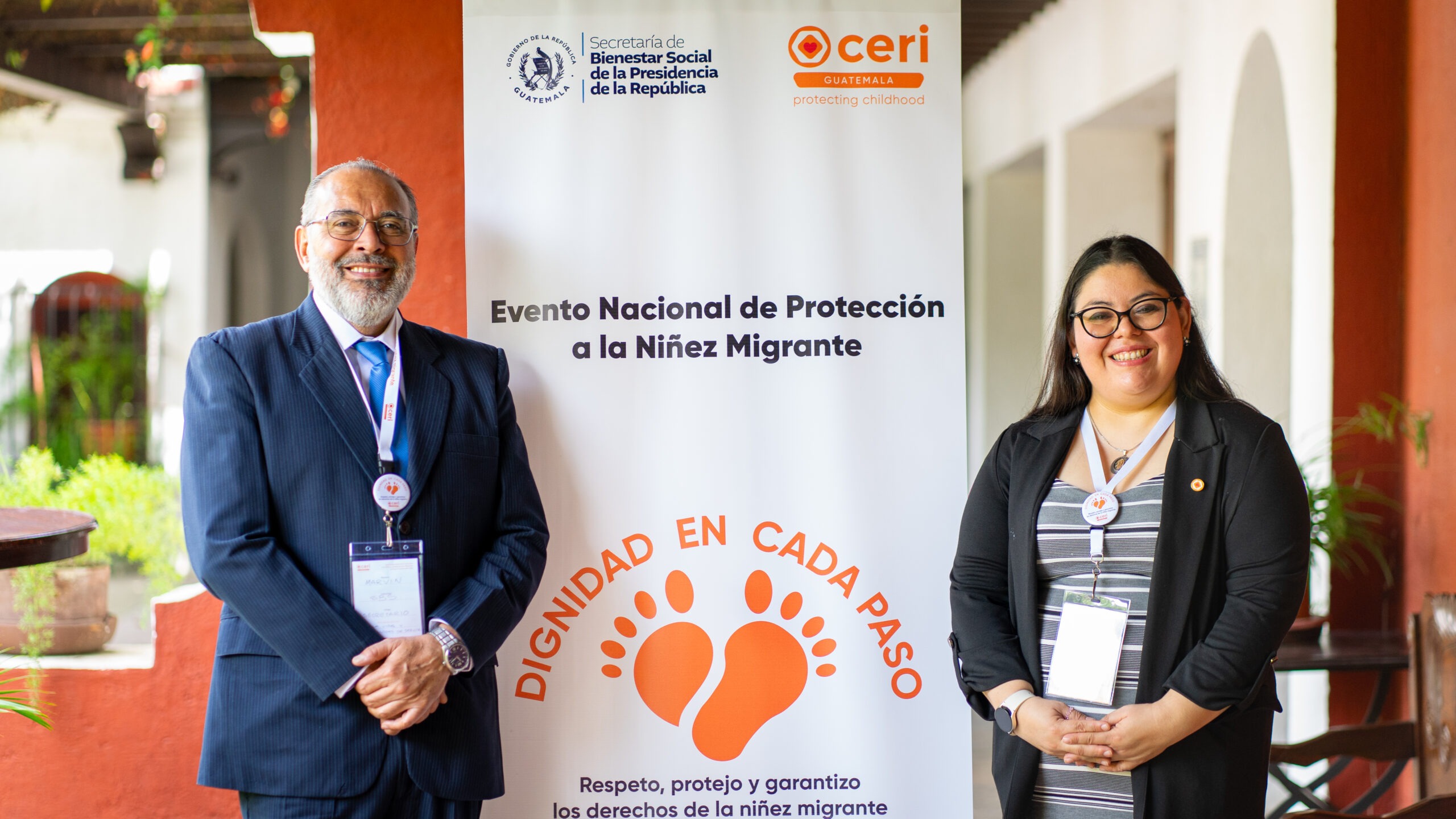 Două persoane, un bărbat în costum și o femeie într-un blazer negru, zâmbesc în timp ce stau în fața unui banner pentru &quot;Evento Nacional de Protección a la Niñez Migrante&quot;, organizat de CERI Guatemala, care promovează demnitatea și drepturile copiilor migranți.